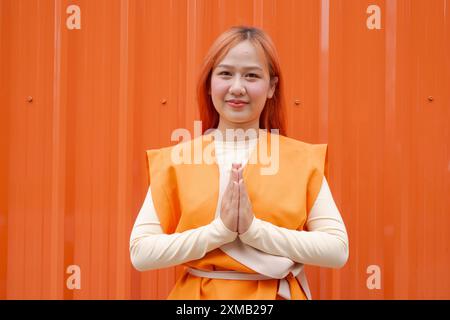 Belle femme thaïlandaise faisant Wai Sawasdee salutation traditionnelle sur fond orange à l'extérieur Banque D'Images