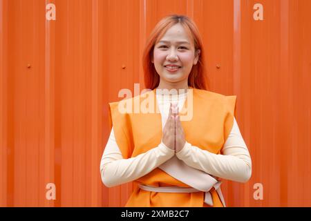 Belle femme thaïlandaise faisant Wai Sawasdee salutation traditionnelle sur fond orange à l'extérieur Banque D'Images