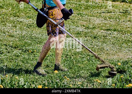 Paysagiste coupe les mauvaises herbes avec un coupe-herbe à ficelle, ses jambes exposées couvertes d'herbe déchiquetée. Banque D'Images