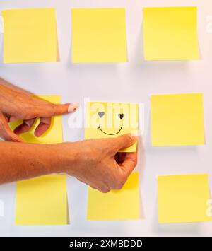 Une personne pointe vers un autocollant jaune de visage souriant sur un tableau blanc. L'autocollant est placé au milieu de la planche, entouré d'autres St jaunes Banque D'Images