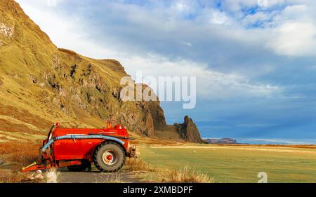 Un véhicule agricole rouge est garé dans un champ à côté d'une montagne. Le ciel est nuageux et le soleil brille à travers les nuages. La scène est paisible et Ser Banque D'Images