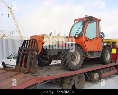 Chariot élévateur orange extra-robuste chargé sur un camion à plateau pour le transport dans une zone de construction. Banque D'Images