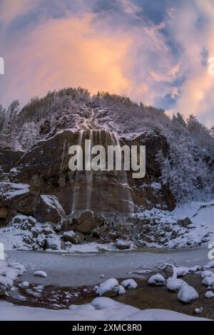 Une impressionnante cascade glacée descend en cascade sur une falaise enneigée, les couleurs chaudes du lever du soleil illuminant le nuage au-dessus. Banque D'Images
