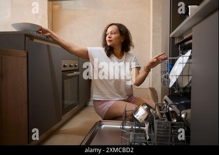 Une femme portant un t-shirt blanc et un short rose est vue en train de vider un lave-vaisselle dans un décor de cuisine contemporain. La scène capture un moment de routi quotidien Banque D'Images