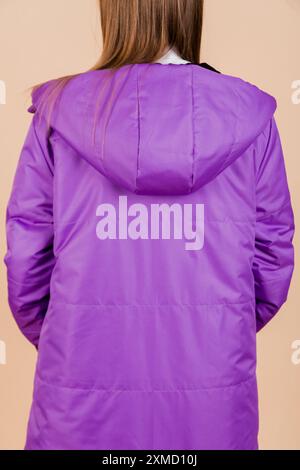 Gros plan détaillé d'une femme portant un manteau d'hiver à capuche violet, photographié de dos sur fond beige. Banque D'Images
