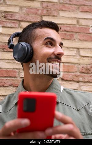 Gros plan jeune homme souriant avec des écouteurs tout en tenant un téléphone portable rouge avec fond de mur de briques Banque D'Images
