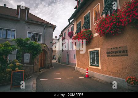 Autriche - 26 octobre 2023 : Une rue étroite dans une ville européenne, avec des bâtiments aux couleurs vives bordés de fleurs rouges. Banque D'Images