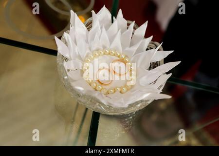 Deux bagues de mariage en or et un collier de perles reposent sur une table en verre transparent, dans un plat en cristal recouvert d'une serviette blanche décorative. Banque D'Images