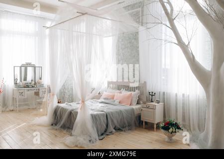 Chambre élégante avec lit blanc avec baldaquin, arbre décoratif, vanité avec miroir, oreillers roses doux, arrangement floral, filtrage de la lumière naturelle Banque D'Images