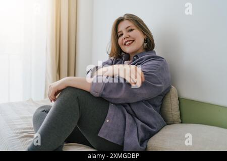 Femme grande taille souriante assise sur le lit, regardant la caméra avec une expression détendue et confiante. Environnement domestique confortable, acceptation de soi et positivité. Style de vie décontracté et confortable. Banque D'Images
