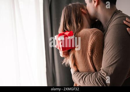 Jeune couple avec boîte cadeau embrassant à la maison profitant de moments romantiques. Célébration de la Saint-Valentin Banque D'Images