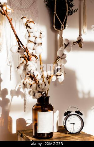 Photo verticale de décoration intérieure confortable avec des tiges de coton dans un vase en verre brun et un réveil noir classique sur une table en bois. Bohème et rustique Banque D'Images