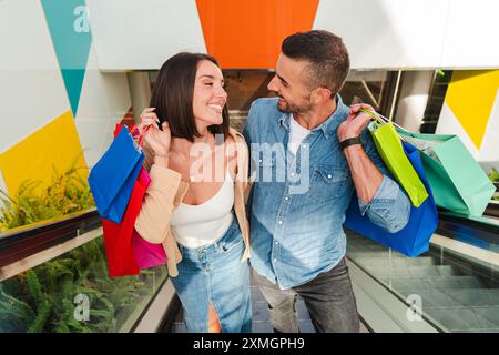 Jeune couple caucasien se regardant dans les yeux tout en tenant des sacs à provisions et en montant l'escalier roulant dans le centre commercial du centre-ville pour aller acheter des bonnes affaires dans un magasin de vêtements. Des gens heureux dans un escalier. Photo de haute qualité Banque D'Images