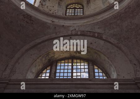 Vue intérieure des fenêtres cintrées dans une église abandonnée, mettant en valeur les murs altérés, la peinture écaillée et la lumière naturelle filtrant à travers le quadrillage Banque D'Images