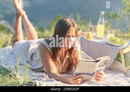 Une femme se détend sur une couverture de pique-nique, lisant un livre avec une table avec des citrons, des raisins et une boisson. Banque D'Images