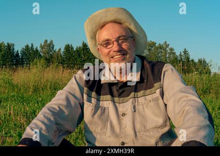 Un homme portant un costume d'apiculture et un chapeau de paille est assis sur l'herbe dans un champ ensoleillé, profitant d'une pause. Le paysage serein et la lumière du soleil brillent Banque D'Images