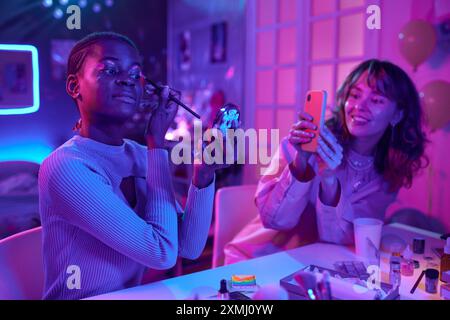 Portrait de deux jeunes femmes utilisant le maquillage luminescent dans la chambre au néon et prenant des photos avec le téléphone Banque D'Images