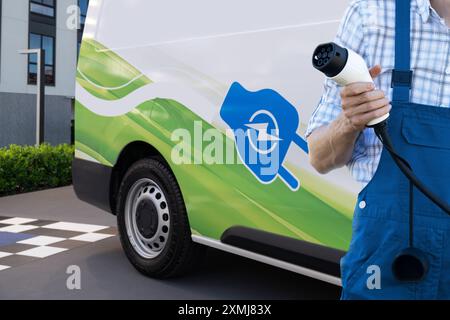 Un homme tient la prise de charge du véhicule électrique. Fourgon électrique en arrière-plan. Banque D'Images