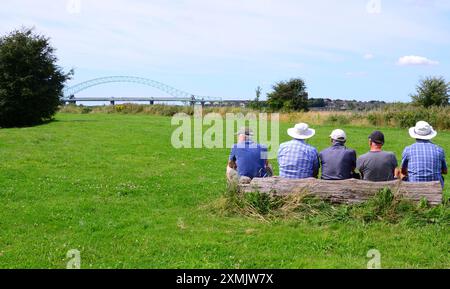 Cheshire, Royaume-Uni, 28 juillet 2024. Juillet se termine chaud dans la région de Widnes Runcorn dans le Cheshire Royaume-Uni, avec une température atteignant 25C. Le dos de cinq hommes assis sur un banc en bois à la campagne. Le pont Silver Jubilee qui relie Widnes à Runcorn est en arrière-plan. Crédit : Terry Waller/Alamy Live News Banque D'Images