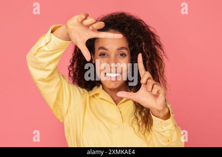 Jeune femme aux cheveux bouclés fait Framing Gesture Against Pink Banque D'Images