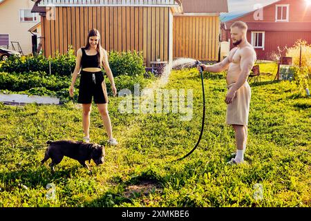 Dans la cour de la maison de campagne, jeune couple marié promène leur chien, qui joue avec des éclaboussures d'eau du tuyau d'arrosage le soir chaud d'été. Banque D'Images