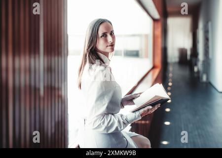 Une femme dans une blouse blanche est assise près d'une grande fenêtre, tenant un livre ouvert. Elle regarde la caméra avec une expression neutre. L'arrière-plan comporte un M. Banque D'Images