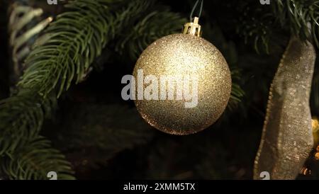 Ornement de Noël scintillant doré et pailleté en gros plan suspendu à un angle dans des feuilles de pin, avec ruban d'or sur le côté. Mise au point sélective. Banque D'Images