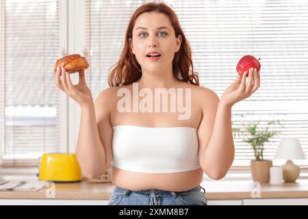 Surpoids jeune femme choquée en jeans serrés choisissant entre le pain et la pomme fraîche à la maison. Concept de gain de poids Banque D'Images