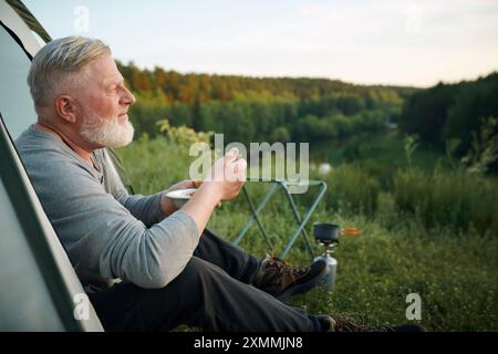 Vue latérale du touriste masculin caucasien senior assis dans la tente de camp manger un repas et profiter du paysage, espace de copie Banque D'Images