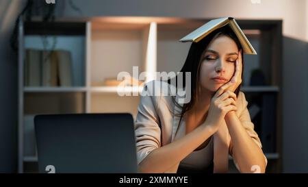 Étudiant fatigué. Travail tardif. Femme épuisée surmenée tenant à la main sa tête avec un livre dessus dormant au bureau préparant l'examen la nuit. Banque D'Images