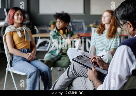 Psychologue masculin mature méconnaissable travaillant avec un groupe d'adolescents prenant des notes sous forme de papier Banque D'Images