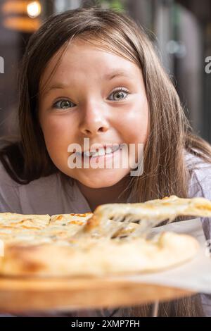 Jolie fille et pizza aux quatre fromages gros plan. Une jeune fille pré-adolescente regarde dans le cadre alors qu'elle se prépare à manger de la pizza. Photo verticale. Foyer sélectionné. Qual. Élevé Banque D'Images