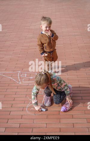 Fille avec son frère cadet griffonnant avec de la craie sur le trottoir, vue de dessus Banque D'Images