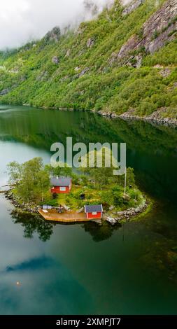 Une vue aérienne d'une petite île boisée dans un fjord norvégien. Deux cabanes rouges sont nichées sur l'île, entourées d'arbres verts et d'un quai en bois. Lovrafjorden, Norvège Banque D'Images