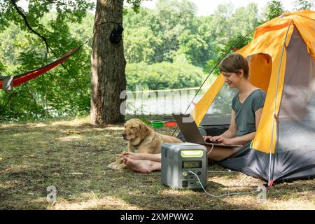 Femme nomade numérique utilise une centrale électrique portable pour charger son ordinateur portable pendant le travail sur la nature sauvage. Son chien allongé à côté. Énergie portable et Banque D'Images