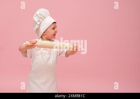 Un enfant habillé en boulanger, avec tablier et chapeau, pose avec un rouleau à pâtisserie en bois sur un fond rose. Enfant âgé de 3 ans (3 ans b Banque D'Images