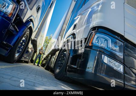 Les camions lourds sont garés étroitement ensemble dans une gare de fret. Un travailleur dans un équipement de sécurité inspecte un des véhicules. Le soleil brille, illumi Banque D'Images