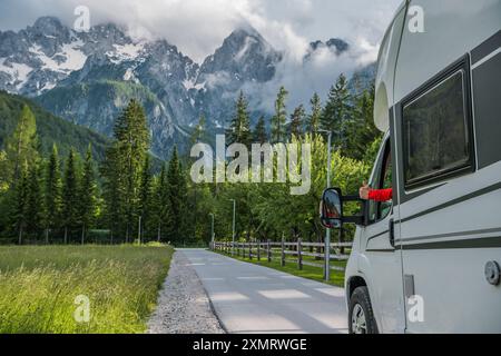 Un camping-car est garé le long d'une route sinueuse qui traverse une prairie verte vibrante, avec des montagnes majestueuses qui s'élèvent au-dessus. Le ciel nuageux ajoute Banque D'Images