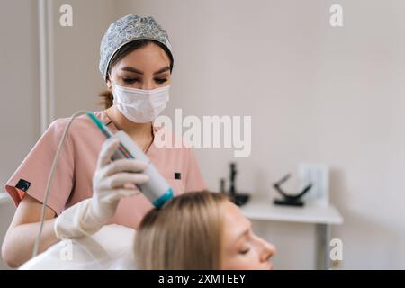 Trichologue qualifié dans les gants et le masque effectuant la procédure de traitement capillaire en utilisant le dispositif de photothérapie de spectre. Patiente blonde ayant des cheveux Banque D'Images