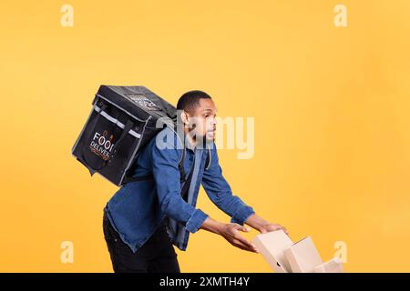 Coursier afro-américain téméraire renversant beaucoup de pizza en studio, laissant tomber une pile massive de boîtes et faisant un désordre. Livreur maladroit et causant un accident, gaspillant de la nourriture. Banque D'Images