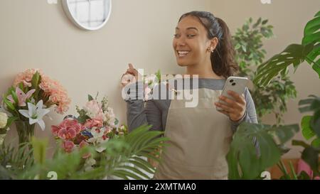 Une jeune femme hispanique souriante portant un tablier beige dans un magasin de fleurs luxuriant, tenant un smartphone tout en se tenant debout parmi diverses plantes en pot et bouque Banque D'Images