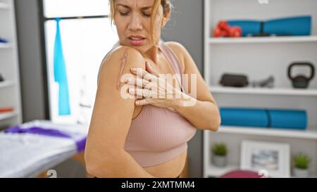 Une femme hispanique examine une cicatrice sur l'épaule dans une salle de clinique de réadaptation, décrivant la récupération et les soins de santé. Banque D'Images