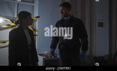 Un homme et une femme, inspecteurs, analysent les preuves dans une scène de crime intérieure faiblement éclairée, entourés d'un ruban de mise en garde. Banque D'Images