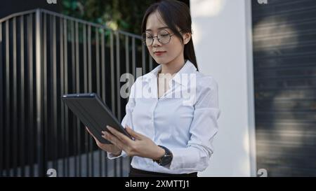 Une jeune femme asiatique en lunettes et en chemise blanche examine attentivement une tablette dans une rue urbaine. Banque D'Images