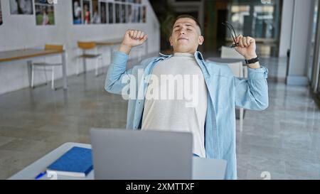 Un jeune homme hispanique s'étire les yeux fermés à côté d'un ordinateur portable dans une bibliothèque universitaire, évoquant un sentiment de soulagement ou de rupture d'études. Banque D'Images
