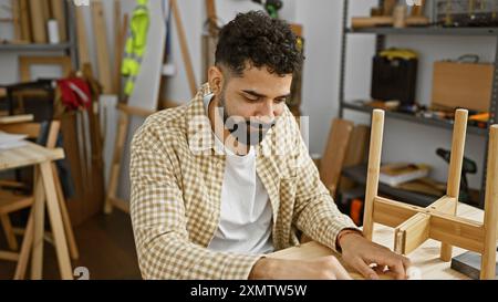 Bel homme artisanal dans un atelier de menuiserie ensoleillé, axé sur l'assemblage de meubles en bois. Banque D'Images