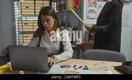 Deux enquêteurs femmes analysent les preuves dans un service de police avec un ordinateur portable, des documents et des photos de crime sur le bureau. Banque D'Images