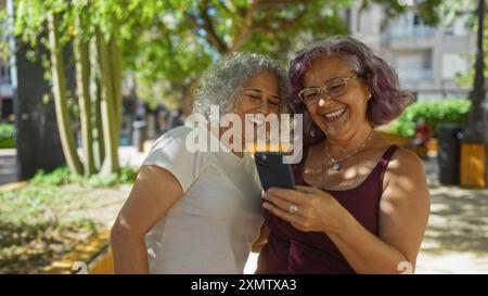 Deux femmes âgées aux coiffures bouclées aiment regarder un smartphone ensemble dans un cadre extérieur pendant une journée ensoleillée. Banque D'Images