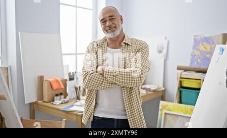 Homme barbu mûr se tient confiant dans un studio d'art entouré de toiles et de fournitures de peinture. Banque D'Images