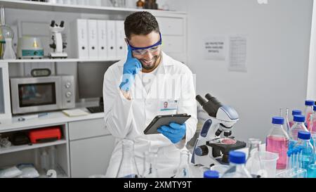 Un homme souriant dans une blouse de laboratoire utilise une tablette numérique dans un laboratoire moderne rempli d'équipements scientifiques. Banque D'Images